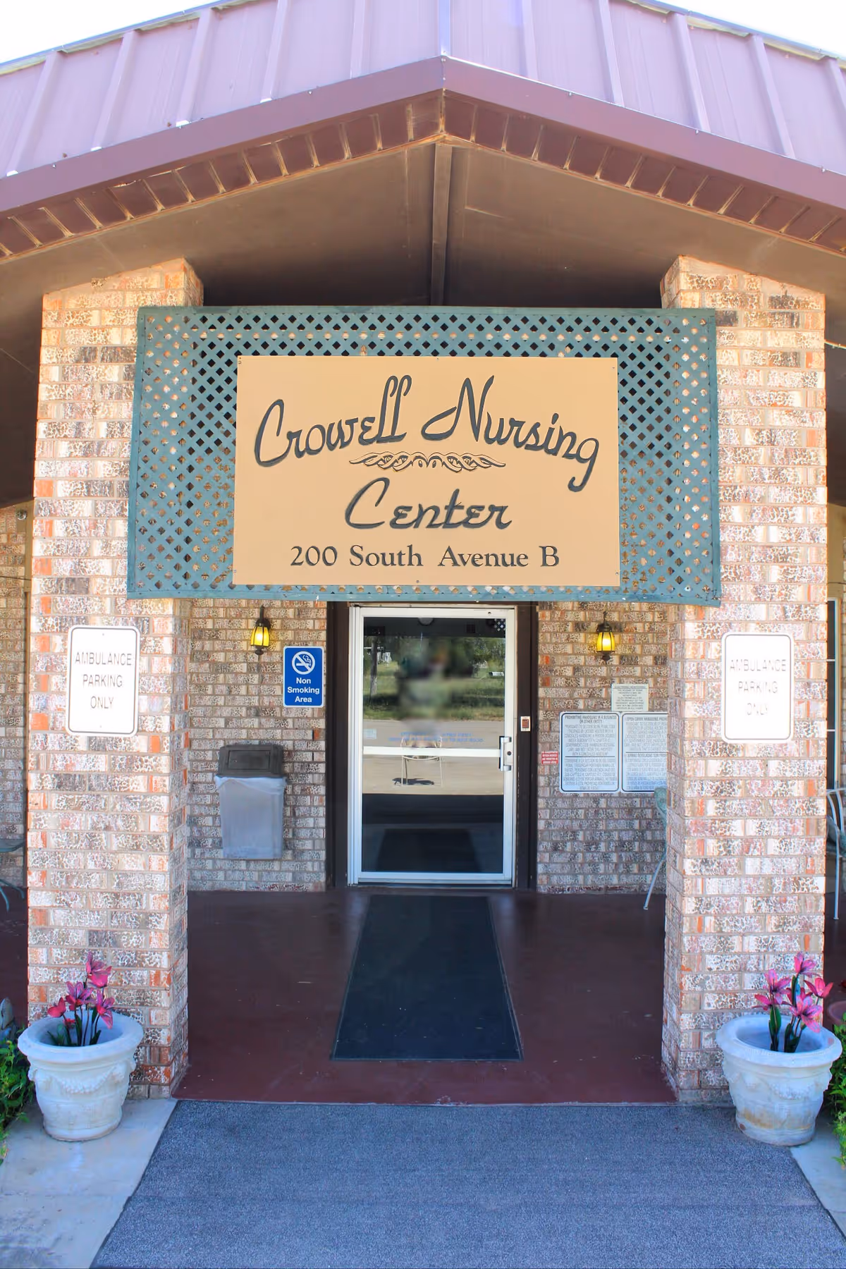Entrance of Crowell Nursing Center with a large sign above the door displaying the facility name and address, flanked by brick pillars and two flower pots with pink flowers on either side of the walkway.