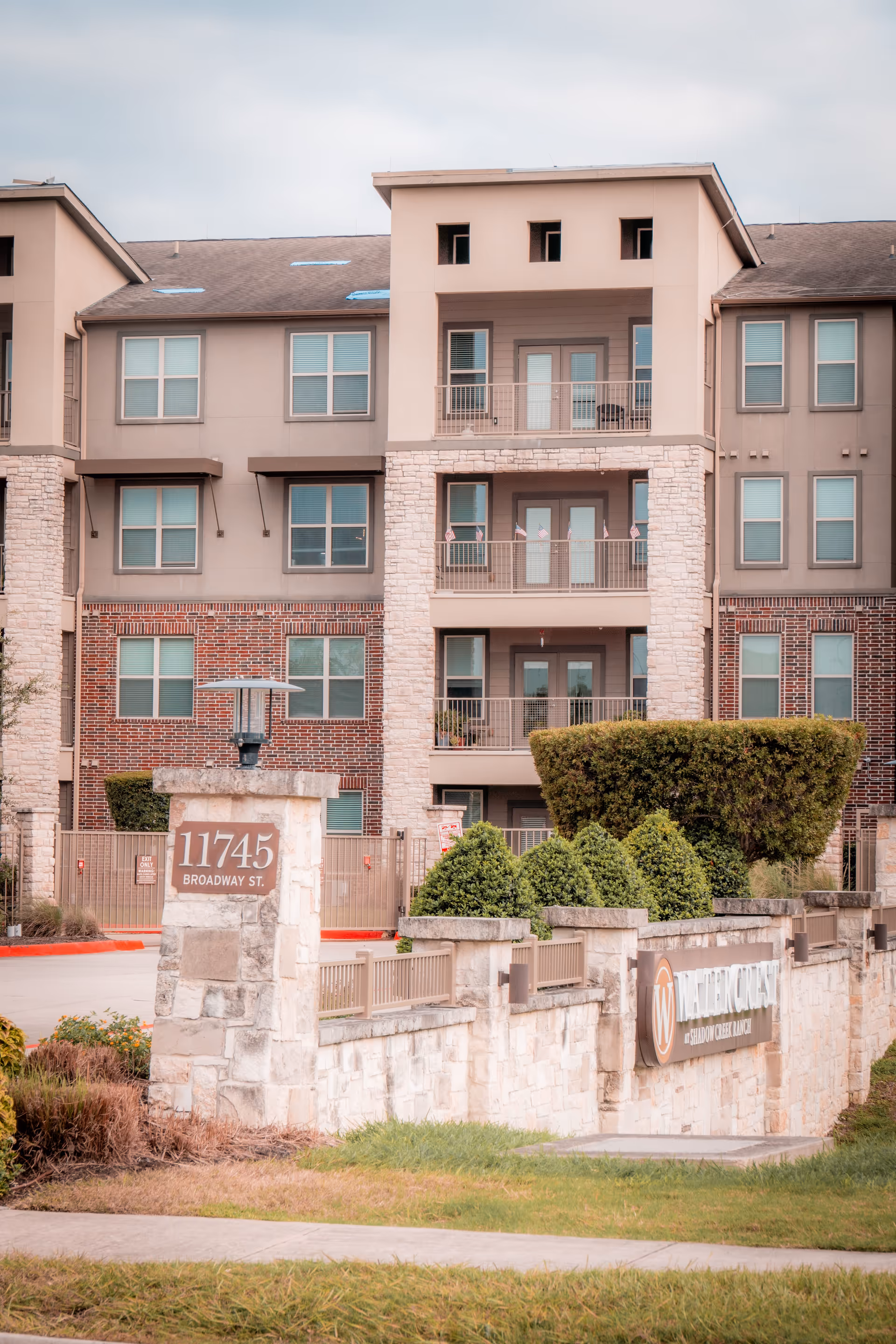 Exterior view of a multi-story senior living facility building with balconies, windows, and a stone entrance sign displaying the address 11745 Broadway St. and the name Watercrest at Shadow Creek Ranch. There are trimmed bushes and a well-maintained lawn in front of the building.