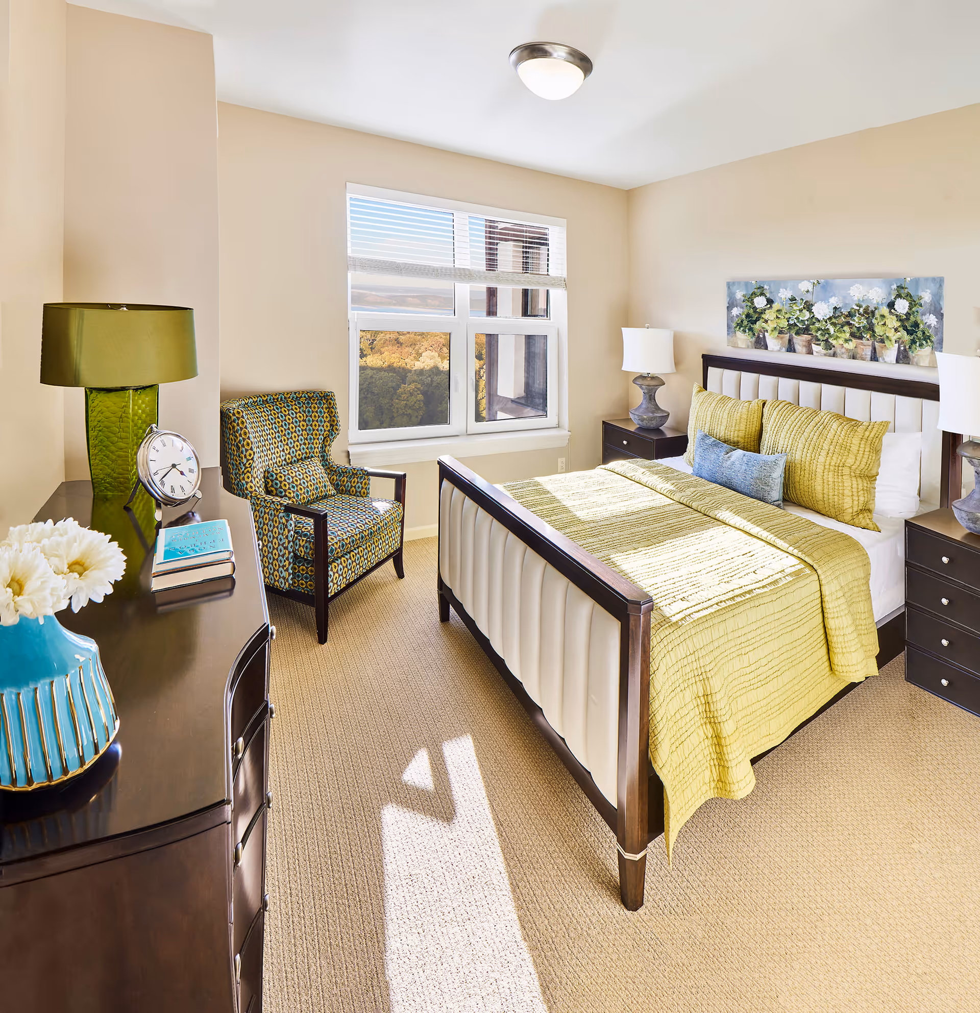 Bright and cozy bedroom with a large window showing an outdoor view. The room features a bed with a green quilt and pillows, two bedside tables with lamps, a patterned armchair, a dresser with a green lamp, clock, books, and a blue vase with white flowers. A floral painting hangs above the bed.