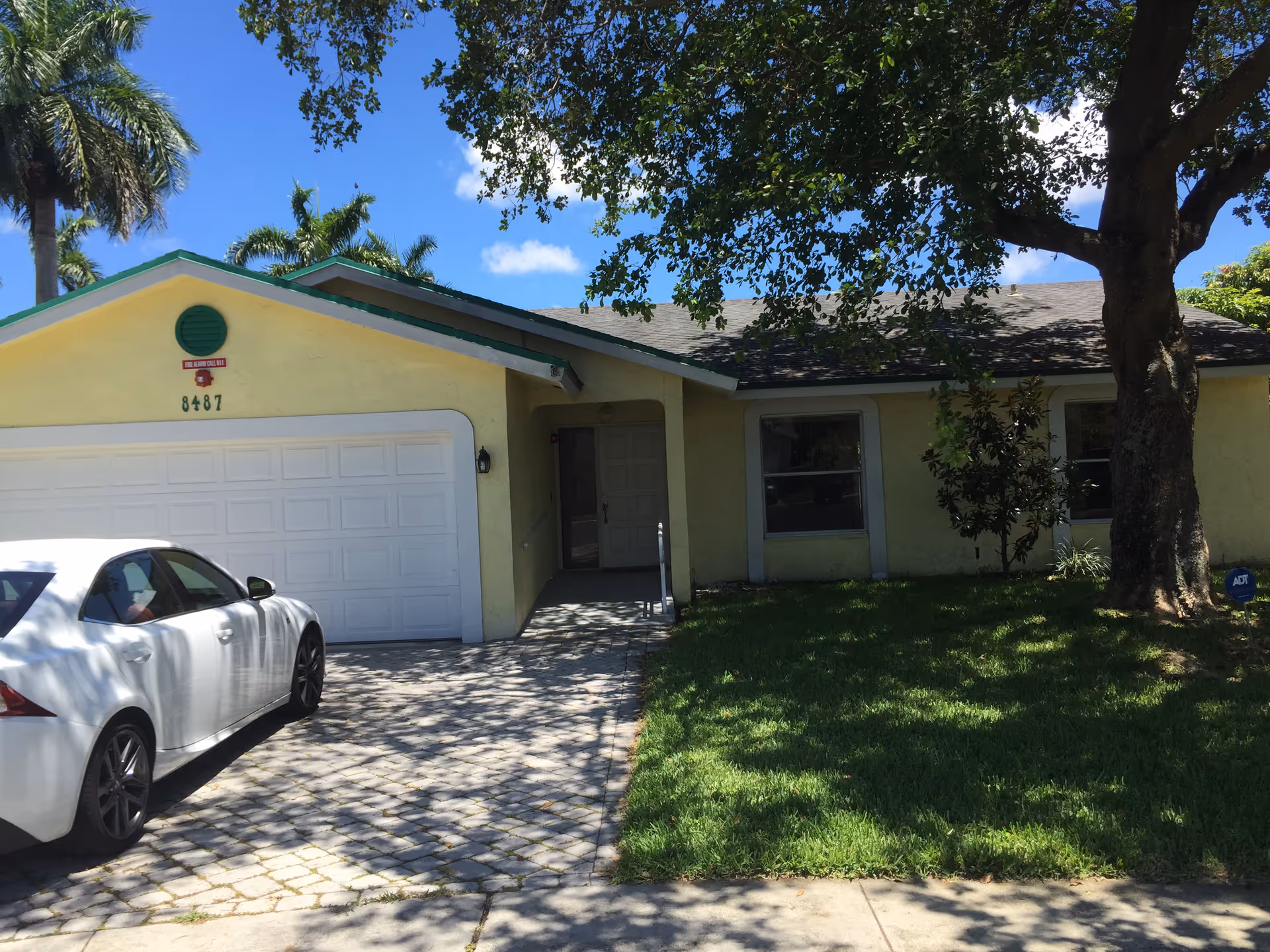 Front exterior view of a single-story yellow house with a white garage door, a white car parked in the driveway, a large tree casting shadows on the lawn, and a clear blue sky with some palm trees in the background.