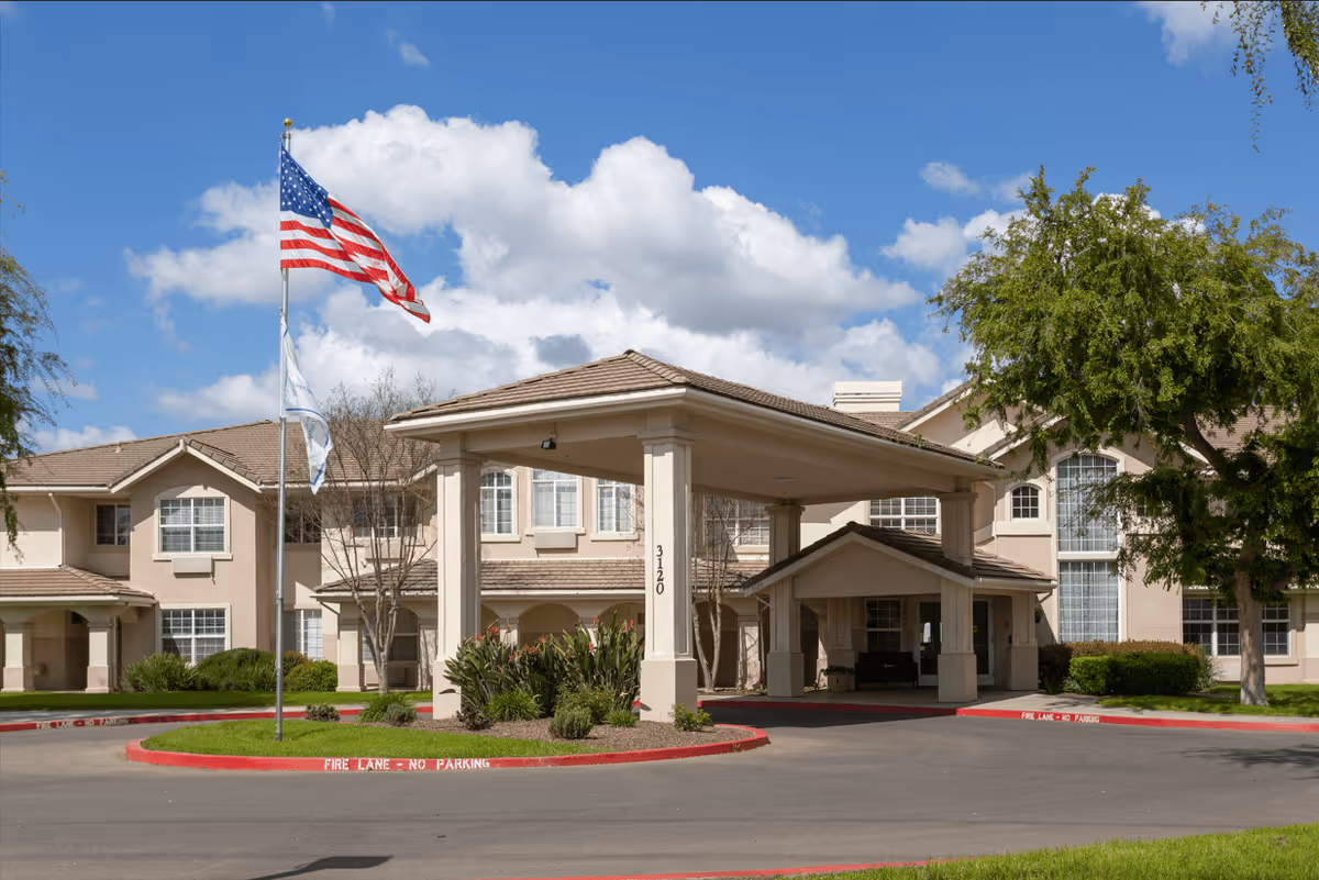 Exterior view of Marbella Visalia senior living facility showing a beige two-story building with large windows, a covered entrance with columns, an American flag and another flag on flagpoles, and landscaping with trees and bushes under a partly cloudy blue sky.