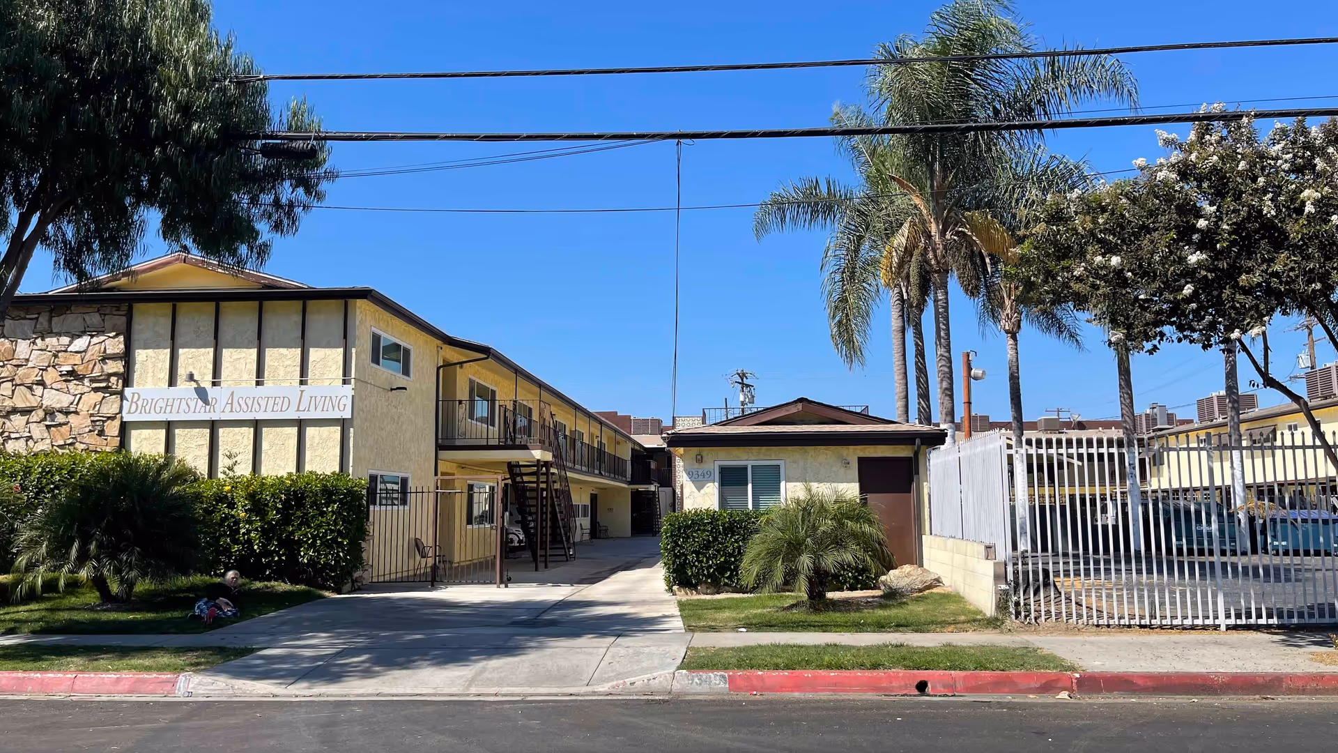 Exterior view of Bright Star Assisted Living facility showing a two-story yellow building with balconies, a gated driveway, palm trees, and a clear blue sky.