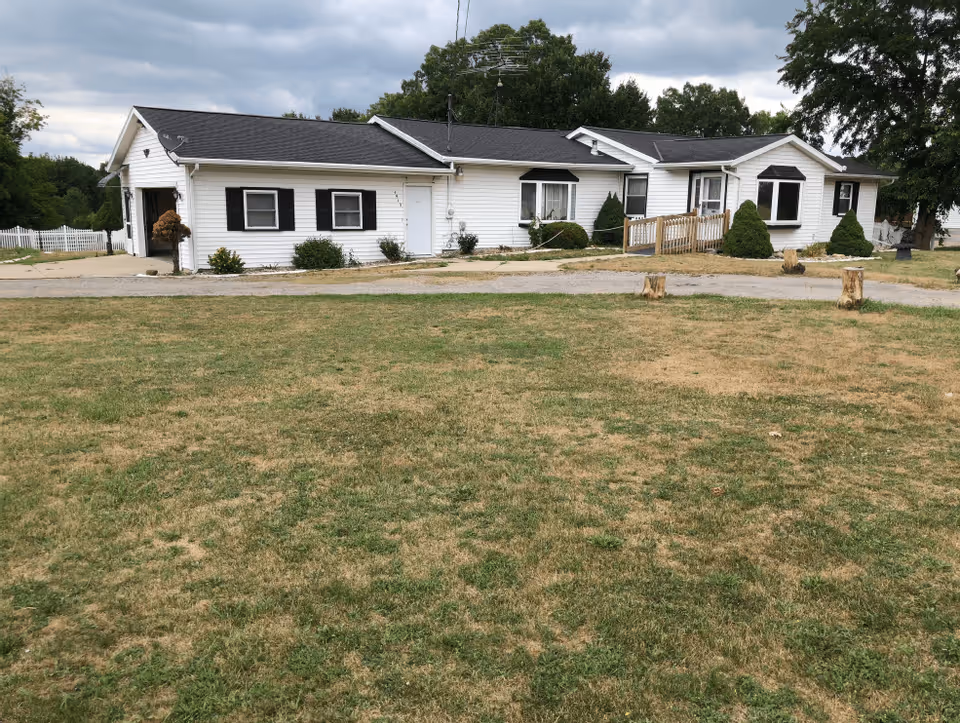 Single-story white building with black shutters and a black roof, surrounded by a grassy yard with some patches of dry grass. There is a paved driveway and a wooden ramp leading to the entrance. Trees and a cloudy sky are visible in the background.