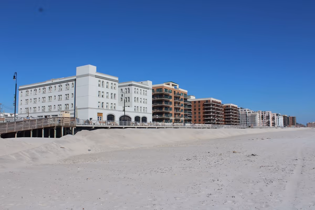 A row of multi-story buildings along a sandy beach under a clear blue sky, with a wooden boardwalk in front of the buildings.