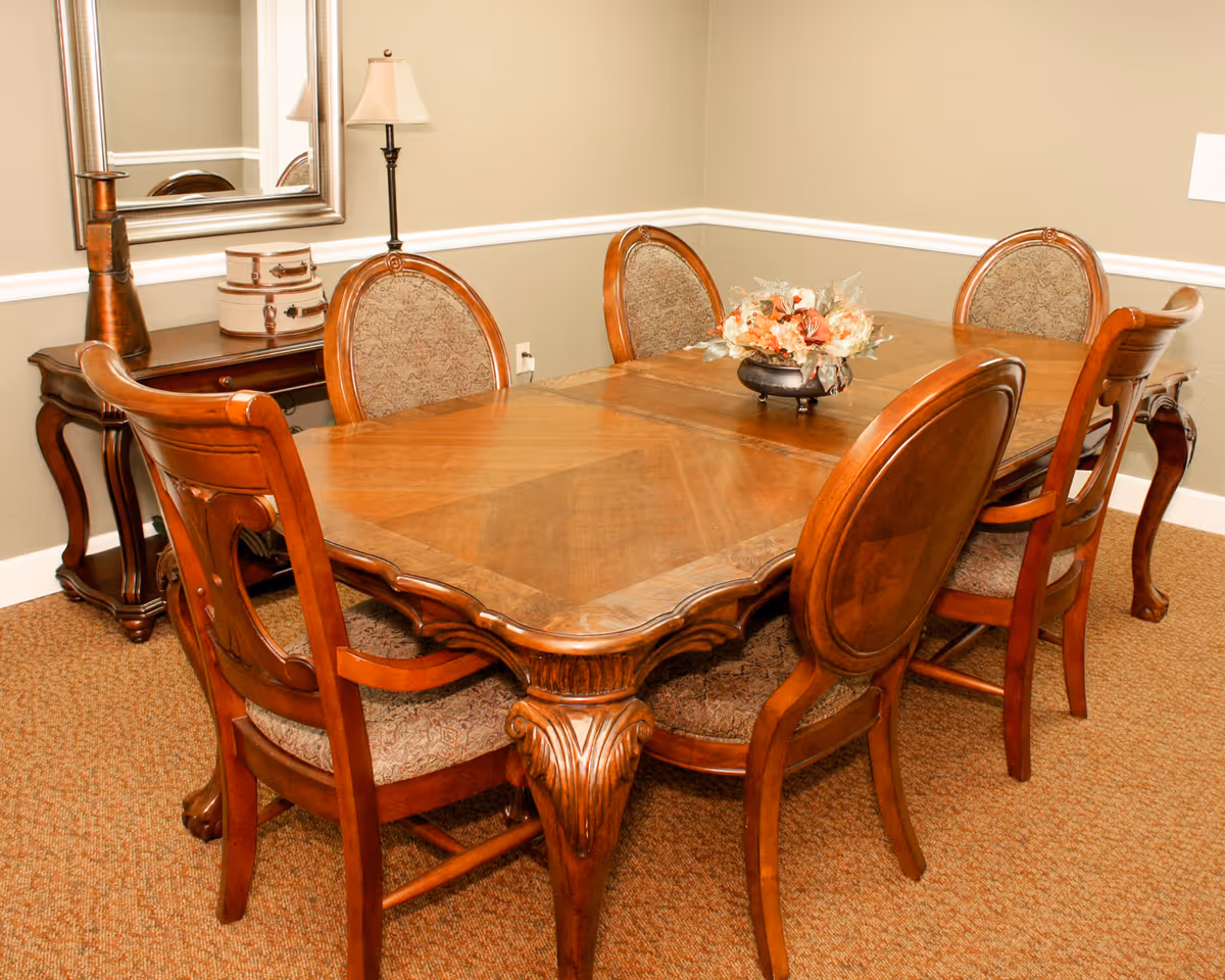 A traditional dining room with a wooden dining table and six matching chairs with upholstered seats and backs. A floral centerpiece is placed on the table. In the background, there is a side table with decorative items and a lamp, and a large mirror hanging on the wall.