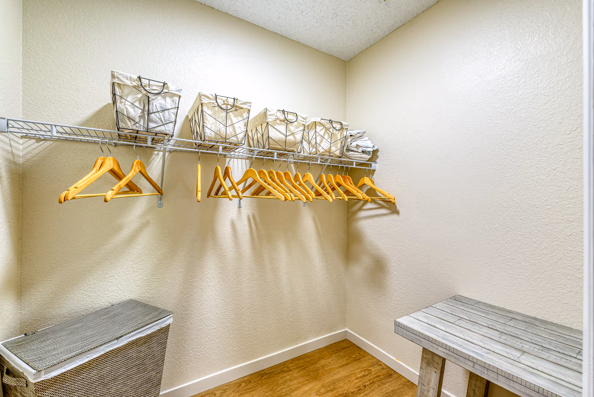 A small walk-in closet with a wooden floor, beige walls, and a white wire shelf mounted on the wall holding several wooden hangers and four fabric storage bins. There is a woven laundry basket on the floor and a small wooden bench on the right side.