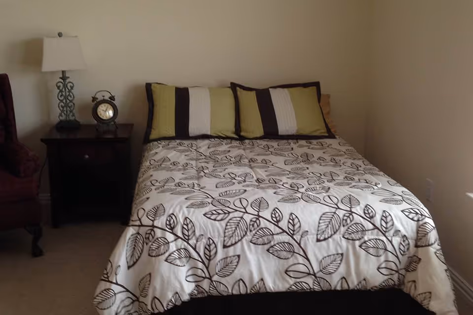 A bedroom with a bed covered in a white comforter with a brown leaf pattern, two pillows with green, white, and brown stripes, a dark wooden nightstand with a decorative lamp and a vintage alarm clock, and a burgundy upholstered chair partially visible on the left.