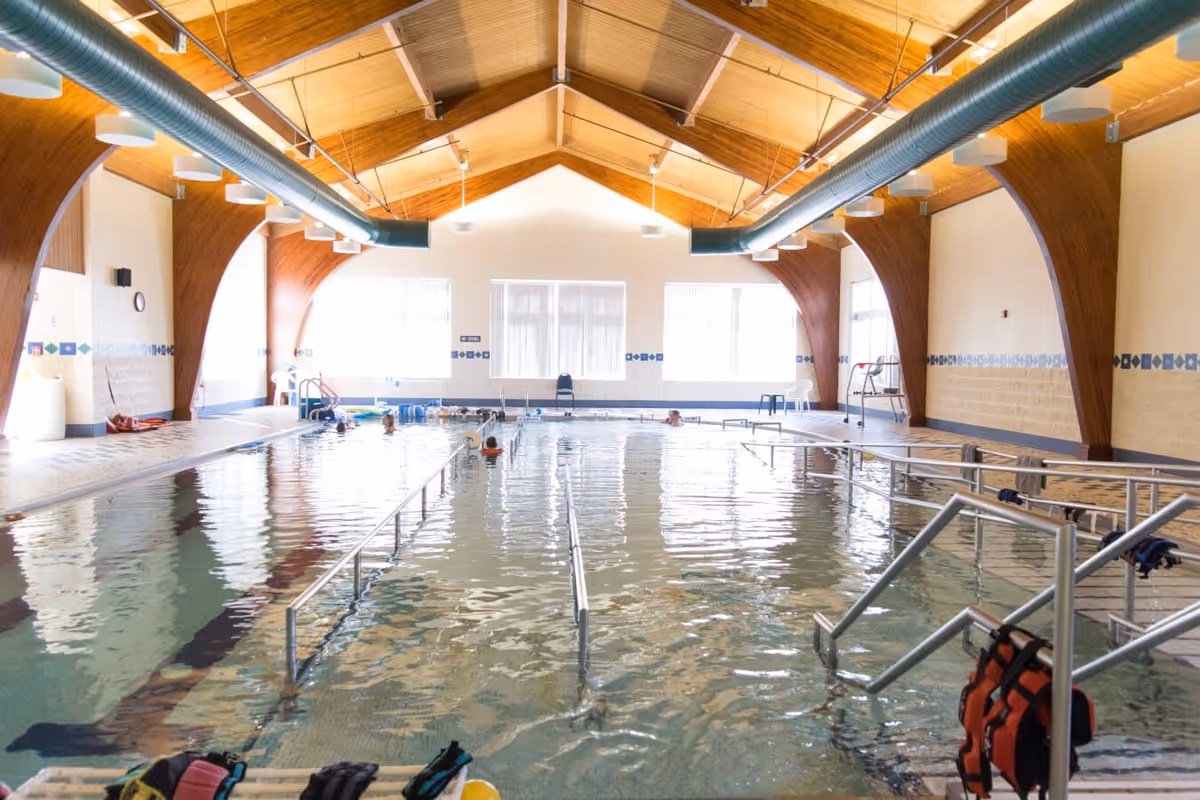 Indoor swimming pool with handrails and steps leading into the water. The pool area has a high wooden ceiling with exposed beams and large windows letting in natural light. Several people are in the pool, and life jackets are hanging on the railing near the steps.