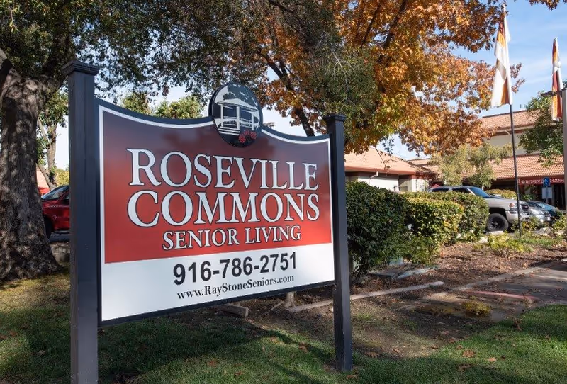 Outdoor view of a large sign for Roseville Commons Senior Living with contact phone number and website, surrounded by trees with autumn leaves and parked cars in the background.