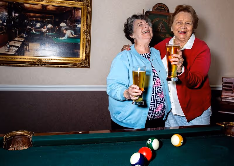 Two elderly women standing next to a pool table, smiling and holding glasses of beer in a room with a framed painting of people playing billiards on the wall behind them.