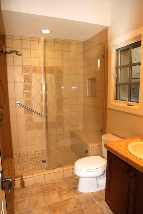 A bathroom with a tiled walk-in shower featuring a glass door and built-in bench, a white toilet, a wooden vanity with a tiled countertop and sink, and a window with a grid design.