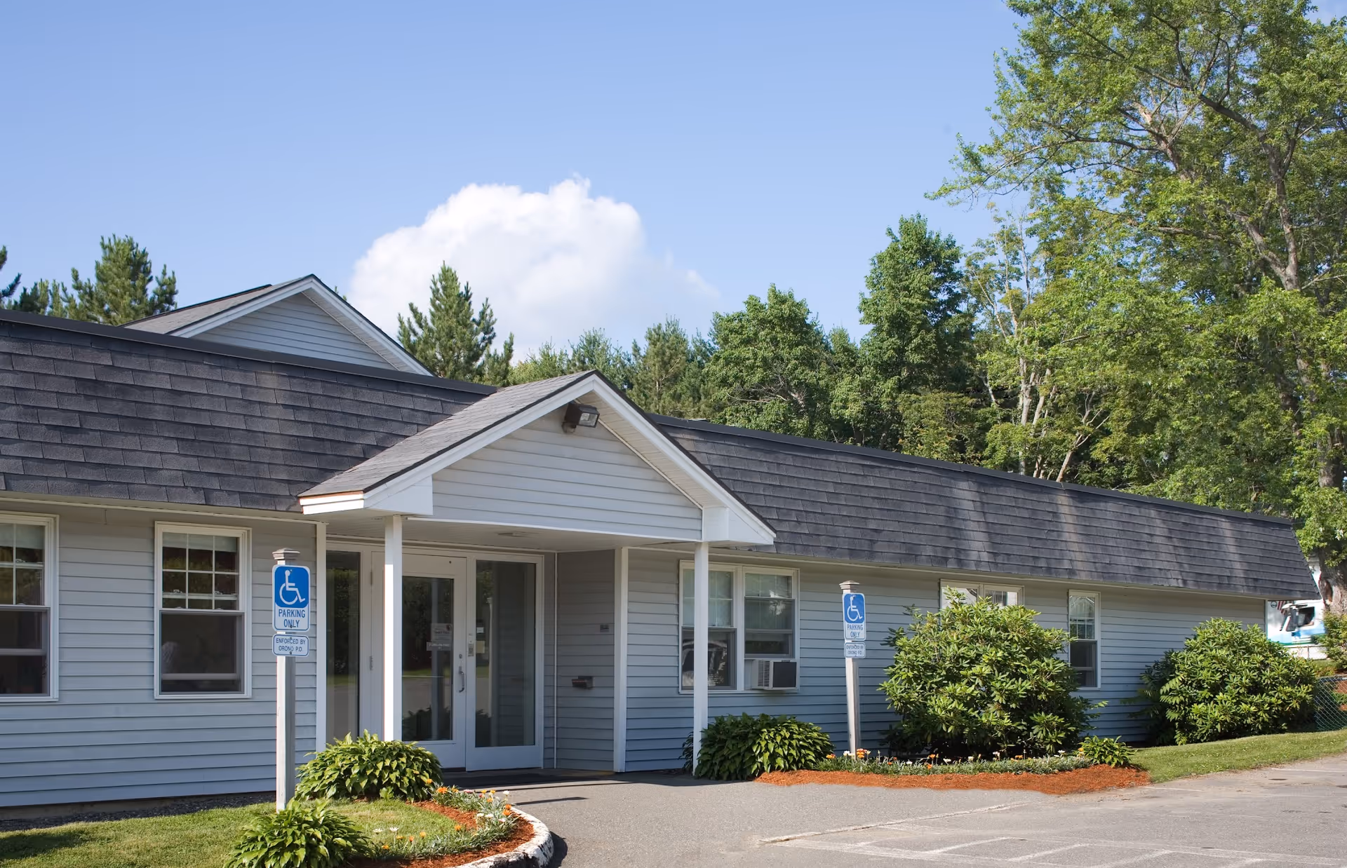 Exterior view of a single-story building with light gray siding and a dark shingled roof under a clear blue sky. The entrance has a small covered porch with double glass doors. There are two handicap parking signs near the entrance, surrounded by green bushes and trees in the background.