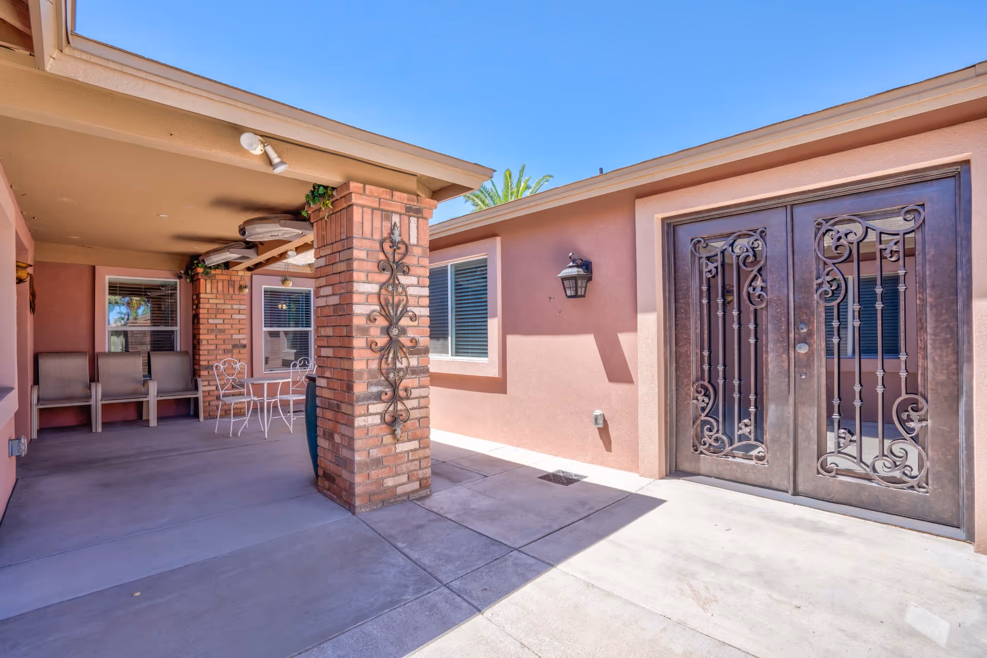 Outdoor covered patio area with brick pillars, metal decorative wall art, a white metal table with chairs, and beige cushioned chairs against the wall. The area is adjacent to a building with a pinkish exterior and large decorative wrought iron double doors under a clear blue sky.