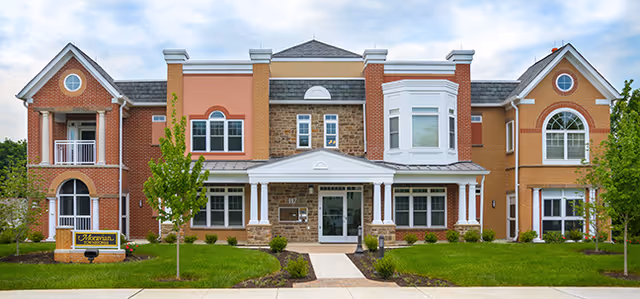 Front exterior view of a two-story senior living facility building with a mix of brick and siding, multiple windows, a central entrance with a covered porch, and landscaped greenery including small trees and shrubs.