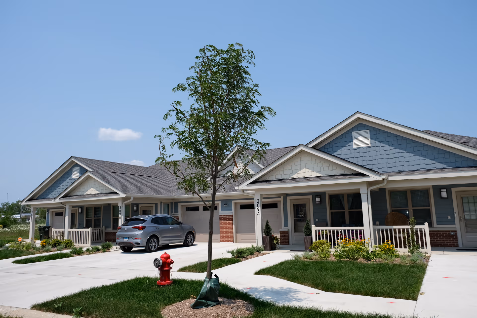 Exterior view of a single-story residential building with blue siding and brick accents. There are two garages, a silver car parked in one driveway, a small tree in the front yard, a red fire hydrant, and a clear blue sky.