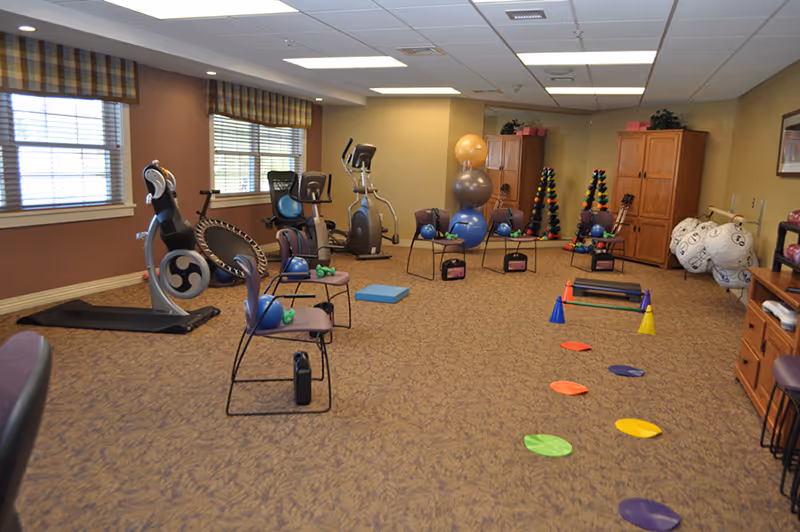A fitness room in a senior living facility with exercise equipment including stationary bikes, an elliptical machine, exercise balls stacked on a chair, dumbbells, and various fitness accessories arranged on the carpeted floor. The room has large windows with blinds and plaid valances, beige walls, and wooden cabinets along the back wall.