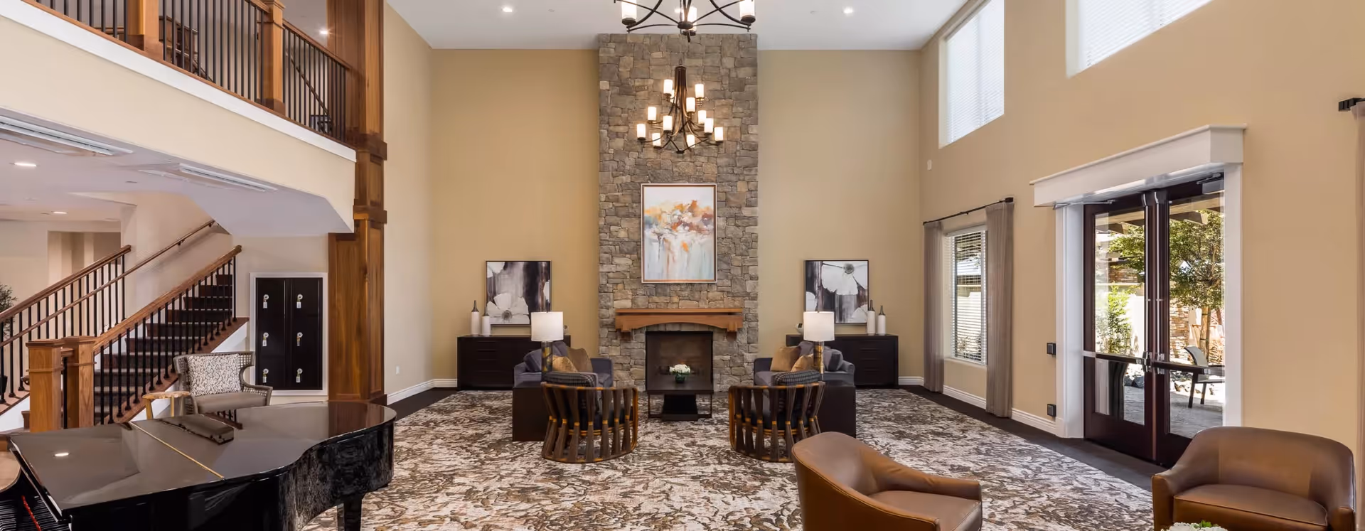A spacious senior living facility common area with high ceilings, a stone fireplace, and large windows. The room features comfortable seating including armchairs and sofas arranged around the fireplace, a grand piano on the left, and a staircase leading to an upper floor. There are decorative paintings on the walls and a chandelier hanging from the ceiling. Double glass doors open to an outdoor patio area.