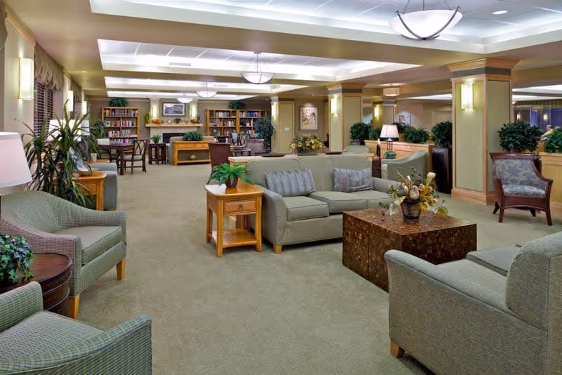A spacious and well-lit senior living facility common area with comfortable seating including sofas and armchairs arranged around a central coffee table. The room features soft lighting, potted plants, and bookshelves in the background, creating a cozy and inviting atmosphere.