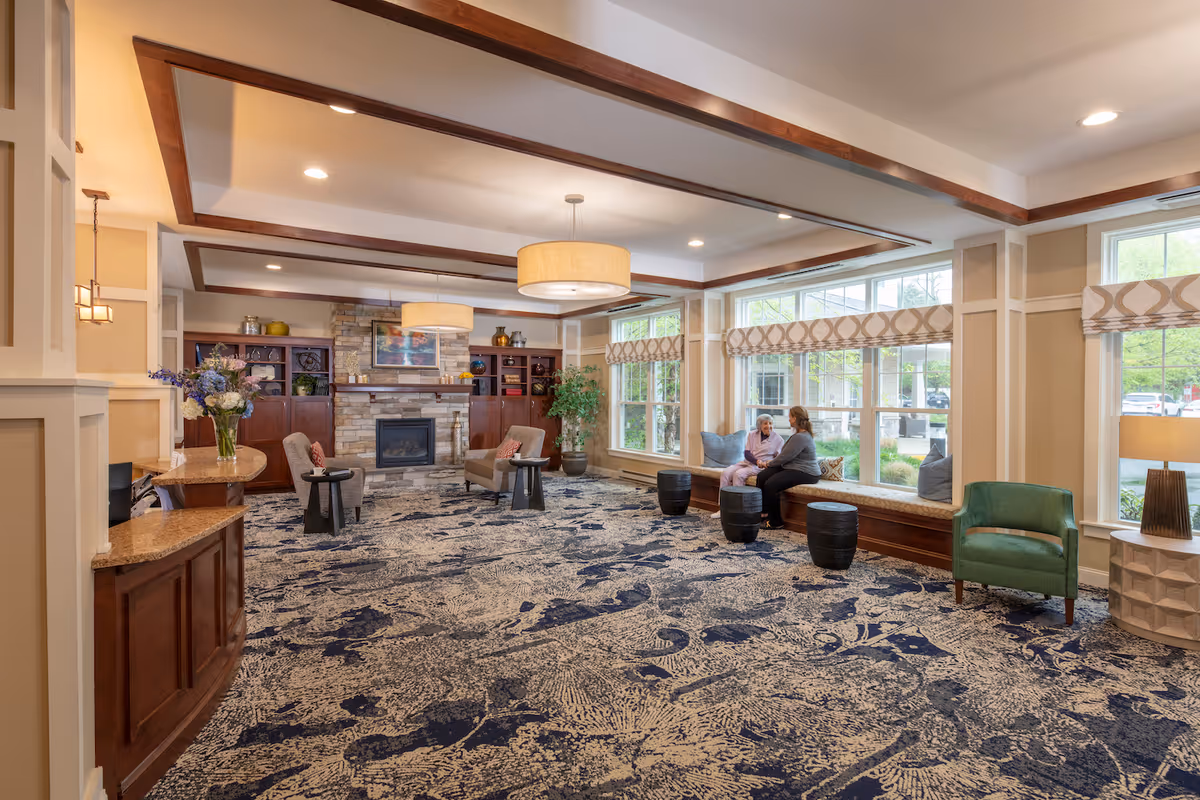 A spacious and well-lit living room area in a senior living facility with large windows, patterned carpet, and comfortable seating including armchairs and window benches. Two people are sitting and conversing near the windows. The room features a stone fireplace, wooden built-in shelves, and modern ceiling lights.