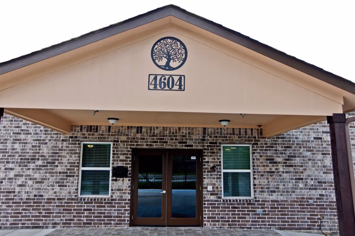 Front exterior view of a brick building with a covered entrance. The building has two windows with blinds on either side of a double glass door. Above the entrance is a beige triangular gable with a tree emblem and the number 4604 displayed below it.