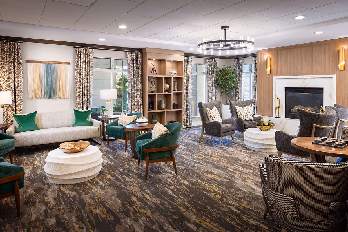 A spacious and well-lit senior living facility lounge with a variety of comfortable seating options including green armchairs, gray wingback chairs, and a beige sofa with green pillows. The room features a modern chandelier, a fireplace with a wooden accent wall, large windows with patterned curtains, a bookshelf with decorative items, and a patterned carpet.