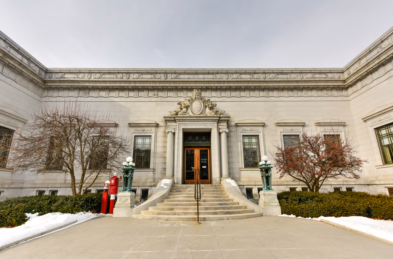 Front exterior view of a classical building with stone steps leading up to a wooden double door entrance. The building features ornate architectural details above the door, flanked by two vintage-style lamp posts. Leafless trees and shrubs are on either side of the entrance, with patches of snow on the ground.