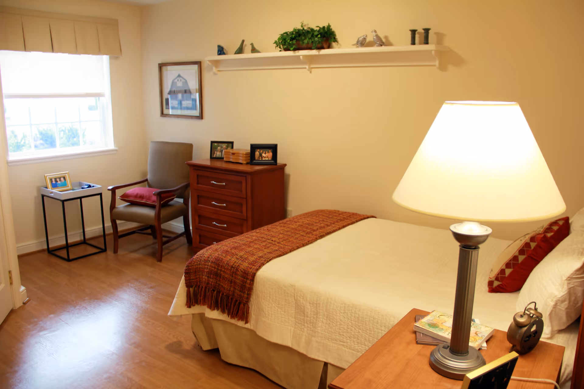 A cozy bedroom in a senior living facility featuring a bed with a beige quilt and red decorative pillows, a wooden nightstand with a lamp and alarm clock, a wooden dresser with framed photos, a comfortable armchair with a red cushion, a small side table with a framed photo, a window with blinds, and a wall shelf with decorative items and a plant.