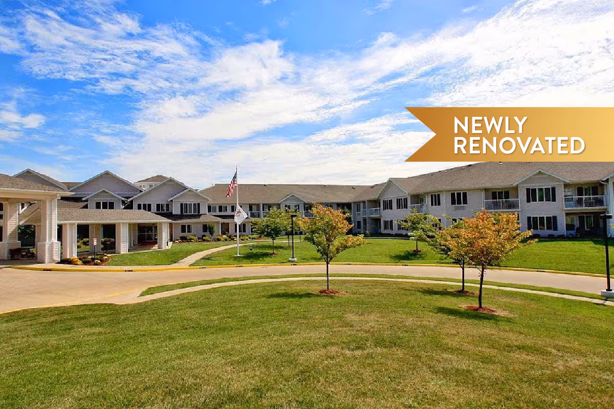 Exterior view of Curtis Creek Senior Living facility showing a large, two-story building with multiple windows and a covered entrance. The building is surrounded by a well-maintained lawn with small trees and a circular driveway. The sky is partly cloudy with blue patches visible. A banner in the top right corner reads 'NEWLY RENOVATED'.