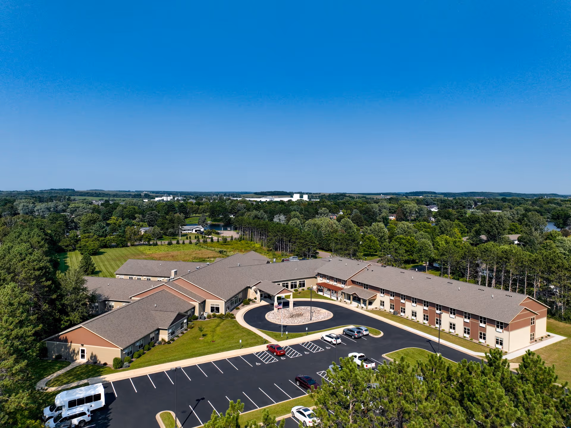 Aerial view of Dove Healthcare - Bloomer facility showing a large, single-story building with a circular driveway and parking lot surrounded by green trees and open fields under a clear blue sky.