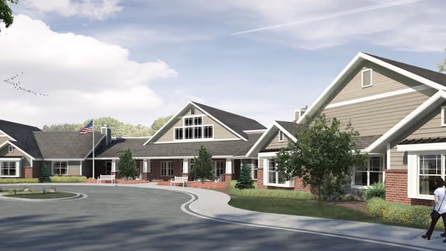 Exterior view of a single-story senior living facility building with beige siding, brick accents, and multiple gabled roofs. There is a curved driveway in front, landscaped greenery, and an American flag on a flagpole. A person is walking on the sidewalk near the building.