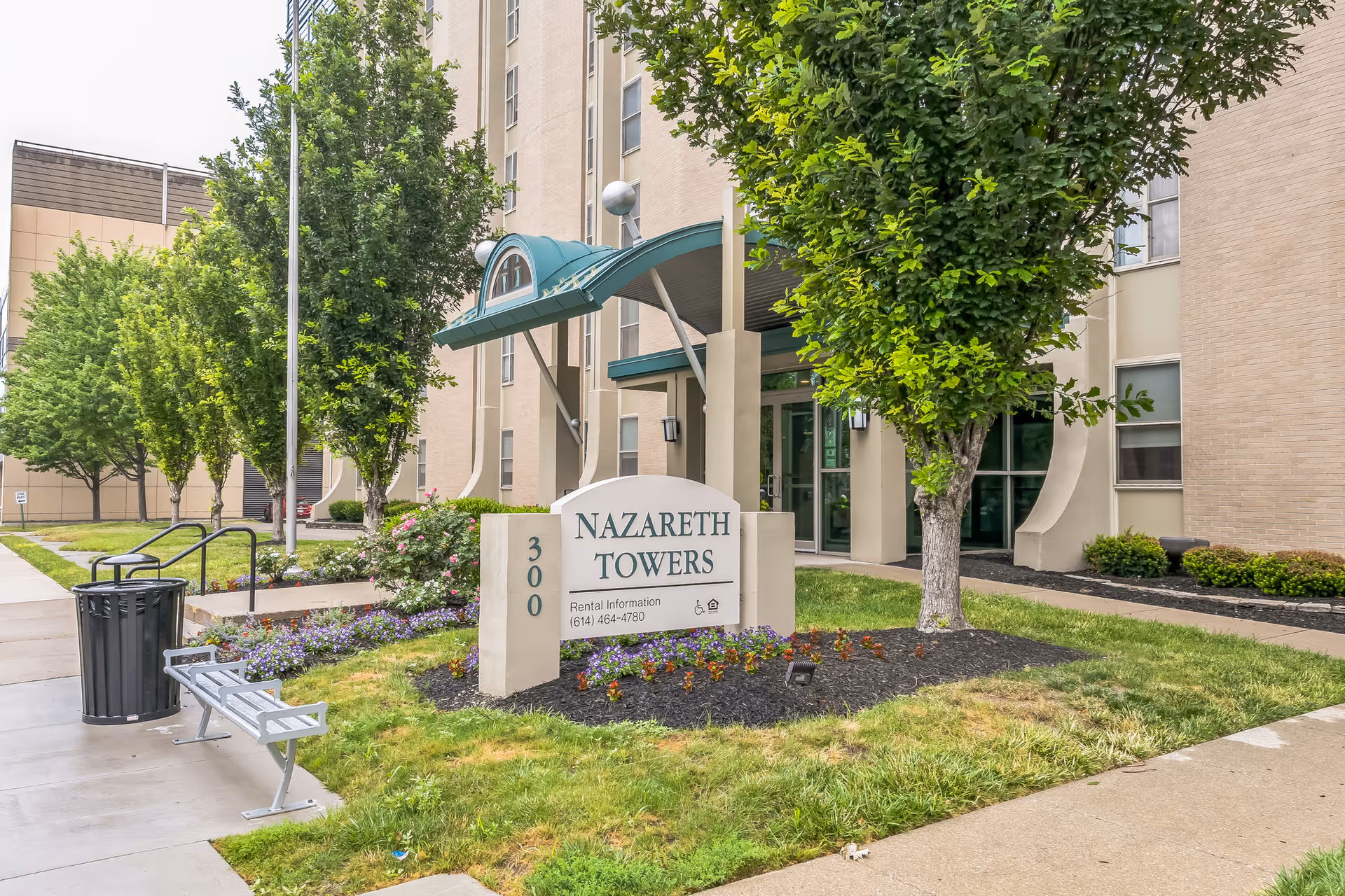 Exterior view of Nazareth Towers building entrance with a sign displaying the name and rental information. The area is landscaped with trees, flowers, and grass, and there is a sidewalk with a bench and trash can nearby.