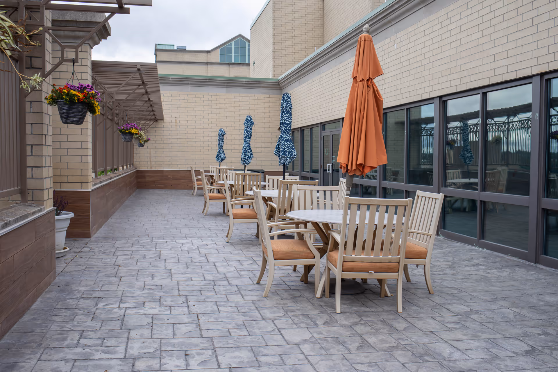 Outdoor patio area with several round tables and chairs. Each table has a closed umbrella, with one orange umbrella in the foreground and several blue patterned umbrellas further back. Hanging flower baskets with colorful flowers are attached to the wall on the left side. The patio is paved with stone tiles and is adjacent to a building with large windows.