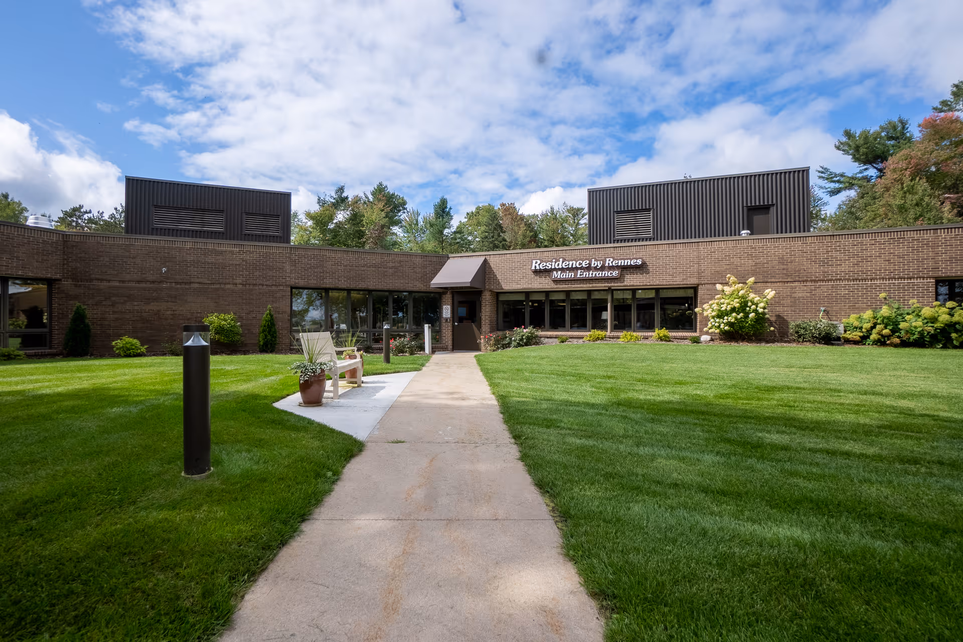 Exterior view of a single-story brick building with a sign that reads 'Residence by Rennes Main Entrance'. A concrete pathway leads to the entrance, flanked by green lawns, potted plants, and a bench. Trees and a partly cloudy sky are visible in the background.