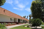 Exterior view of a single-story building with a tiled roof and white walls, surrounded by green grass, shrubs, and trees under a blue sky with some clouds.