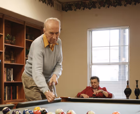An elderly man in a gray sweater and beige pants is playing pool, aiming to hit the cue ball on a billiards table. In the background, an elderly woman in a red top is sitting and watching near a window with blinds. The room has a bookshelf and decorative items on a shelf above the window.