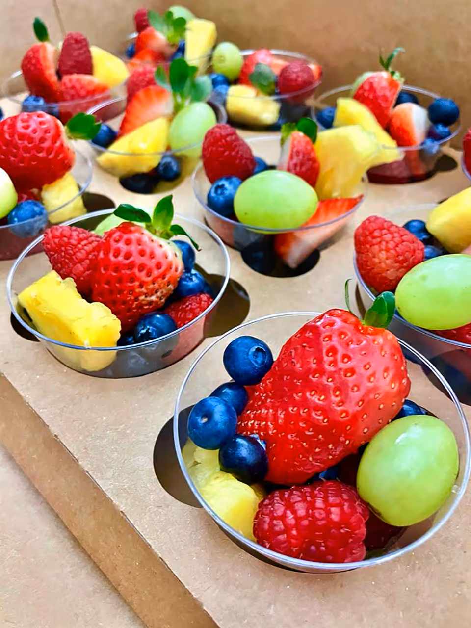 Close-up view of small clear cups filled with a colorful assortment of fresh fruit including strawberries, blueberries, raspberries, green grapes, and pineapple chunks arranged in a cardboard tray.