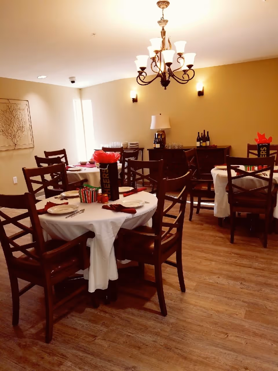 A dining room with round tables covered with white tablecloths, set with plates, utensils, and red napkins. Each table has a centerpiece with red tissue paper and a container labeled 'CHEERS'. Wooden chairs surround the tables. In the background, there is a sideboard with bottles of wine, glasses, and a lamp. The room has warm lighting from a chandelier and wall sconces, with wooden flooring and beige walls.