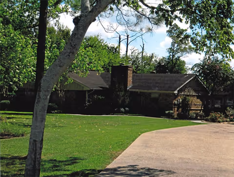 A single-story brick building with a dark roof surrounded by green trees and a well-maintained lawn under a partly cloudy sky. A paved driveway curves in front of the building.