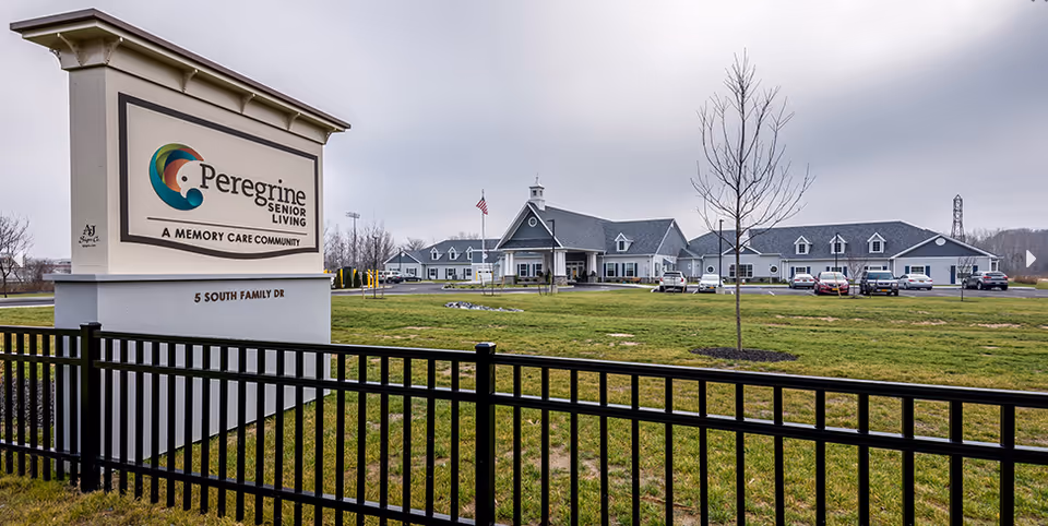 Exterior view of Peregrine Senior Living at Onondaga Hill, a memory care community, showing a large sign with the facility name and address, a black metal fence, a grassy lawn, a few leafless trees, and a long building with multiple windows and a peaked roof under a cloudy sky.