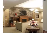 A cozy living room area in a senior living facility featuring a stone fireplace with a mounted flat-screen TV above it, a wooden mantel decorated with a red flower arrangement, a beige sofa, a wooden side table with a floral centerpiece, and soft overhead lighting.