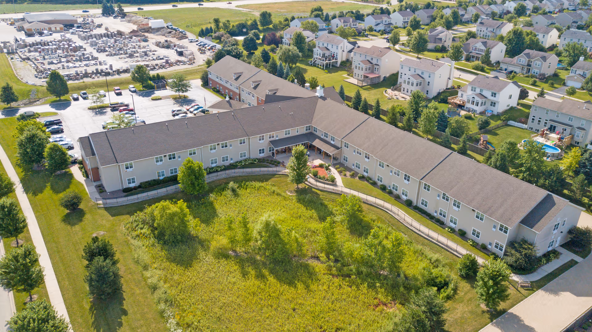 Aerial view of a large senior living facility building with a brown roof surrounded by green lawns, trees, and a walking path. The building is situated near a residential neighborhood with multiple houses and a parking lot with several cars.