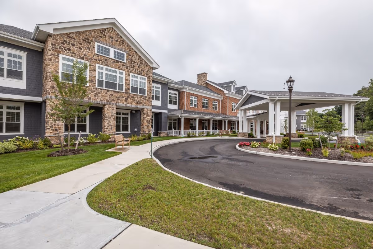 Exterior view of Sunrise of Huntington senior living facility showing a large building with stone and brick facade, multiple windows, a covered entrance with white pillars, a curved driveway, landscaped grass, trees, and flower beds under an overcast sky.