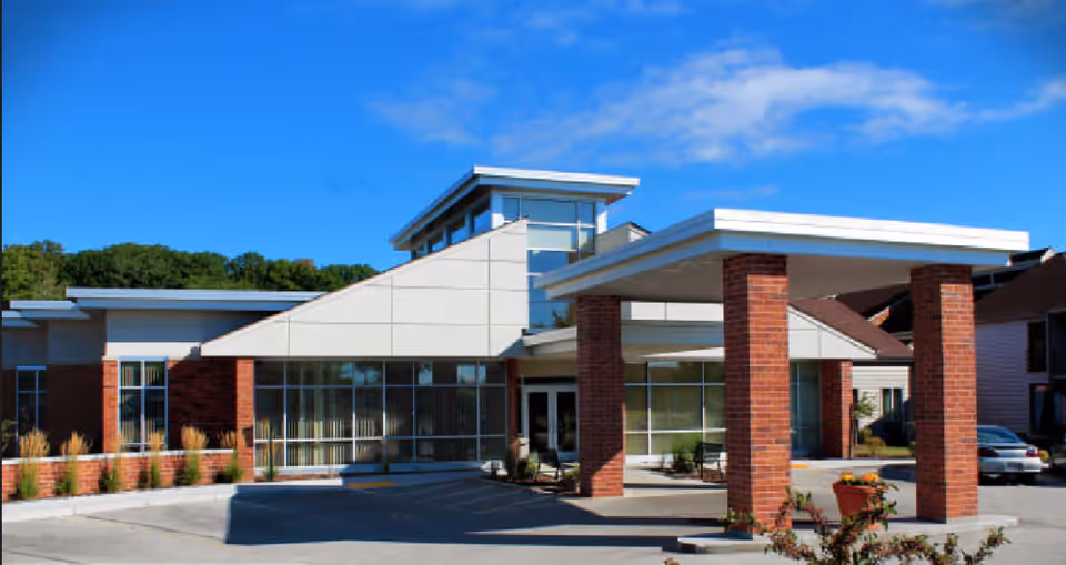 Exterior view of a modern senior living facility building with large windows, brick pillars supporting a covered entrance, and a clear blue sky in the background.
