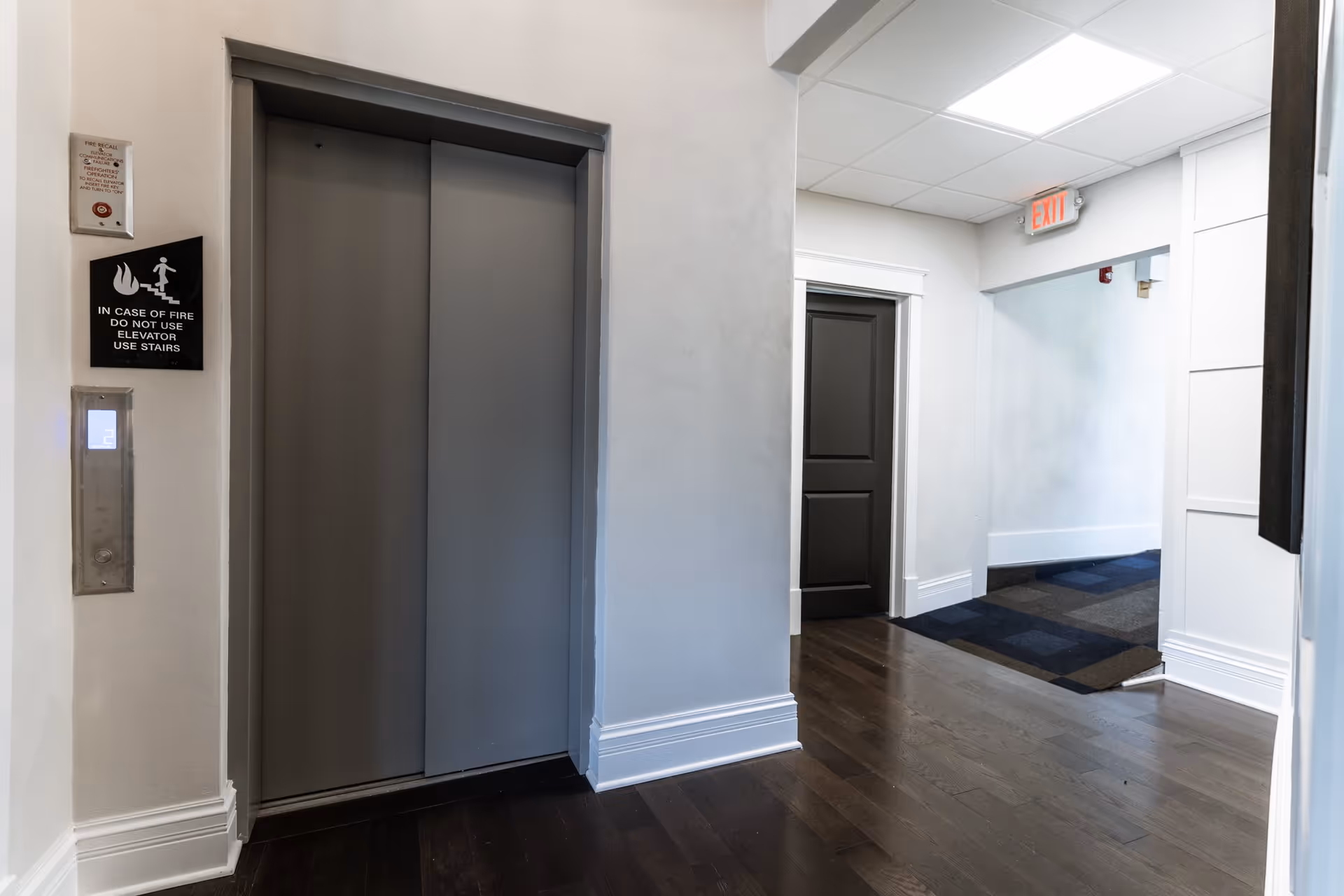 Interior hallway of Elm Haven Place showing a closed gray elevator door with a digital floor indicator displaying the number 2. To the right, there is a dark-colored door and an exit sign above a hallway entrance. The floor is dark wood, and the walls are painted white with some molding details.