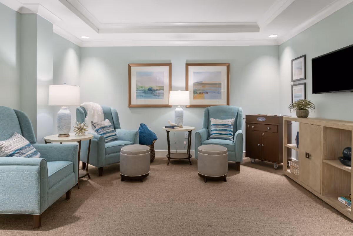 A cozy living room with light blue armchairs, each with a decorative pillow, two small round ottomans, and two side tables with lamps. The walls are painted light green and decorated with framed artwork. There is a wooden cabinet with shelves and a flat-screen TV mounted on the wall above it.