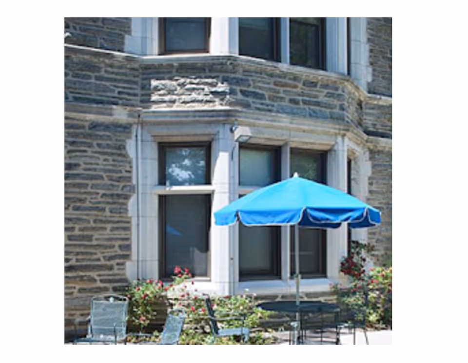 Outdoor seating area with a blue umbrella, metal chairs, and a table in front of a stone building with large windows and some flowering plants.