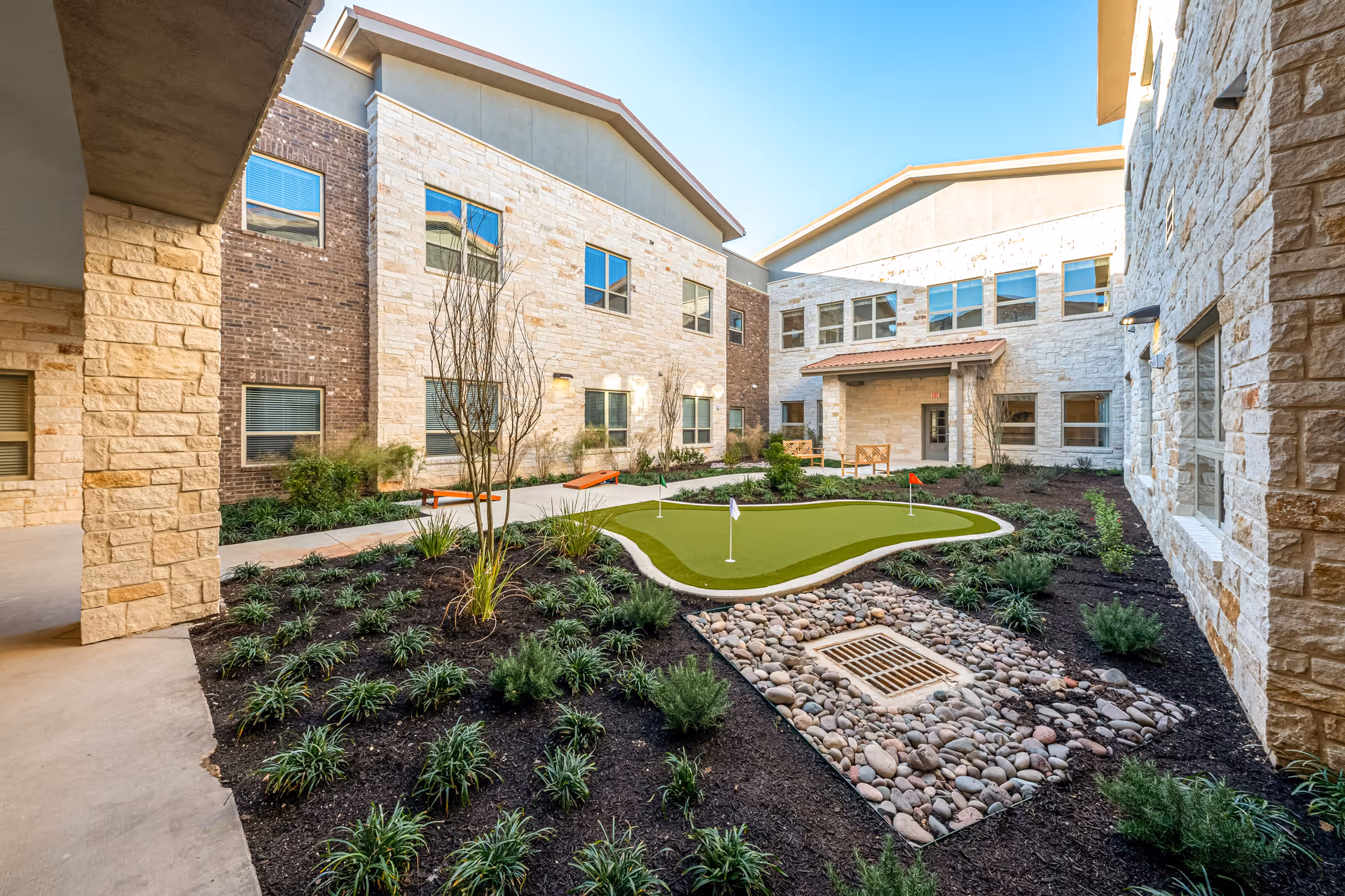 Outdoor courtyard area at Buda Oaks Assisted Living and Memory Care featuring a small putting green with flags, surrounded by landscaped garden beds with plants and shrubs, and a stone and brick building with multiple windows in the background under a clear blue sky.