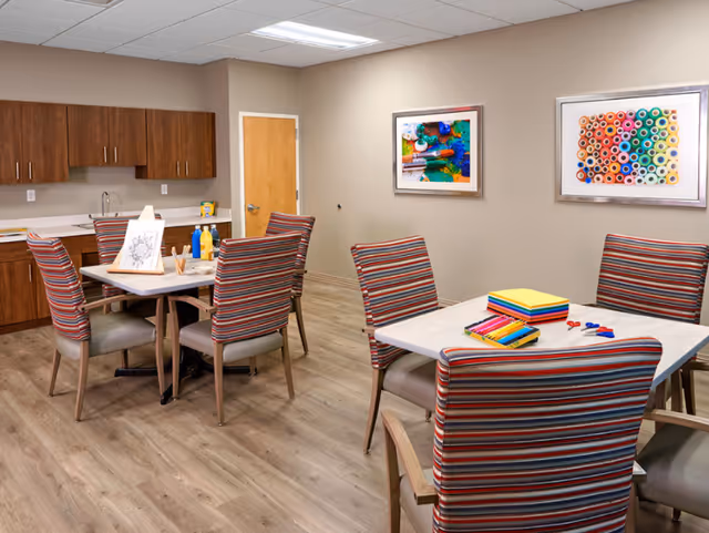 A well-lit room with two tables surrounded by striped upholstered chairs. On the tables are art supplies including colored pencils, scissors, and paper. The room has wood flooring, beige walls, and wooden cabinets with a sink along one wall. Two colorful framed pictures hang on the wall.