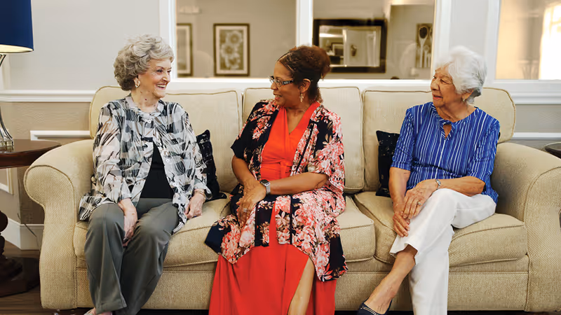 Three elderly women sitting on a beige couch in a well-lit living room, engaged in conversation and smiling. The room features framed artwork on the wall and a side table with a lamp.