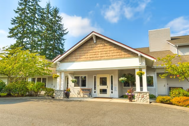 Front exterior view of a senior living facility building with a peaked roof, white siding, stone pillars, hanging flower baskets, and a bench near the entrance under a clear blue sky.